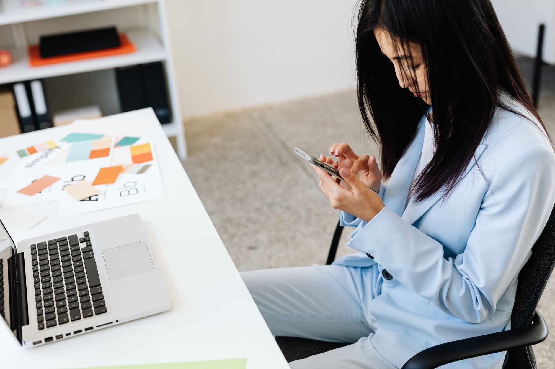 Asian woman in a blue suit using smartphone at office desk with laptop and color samples.