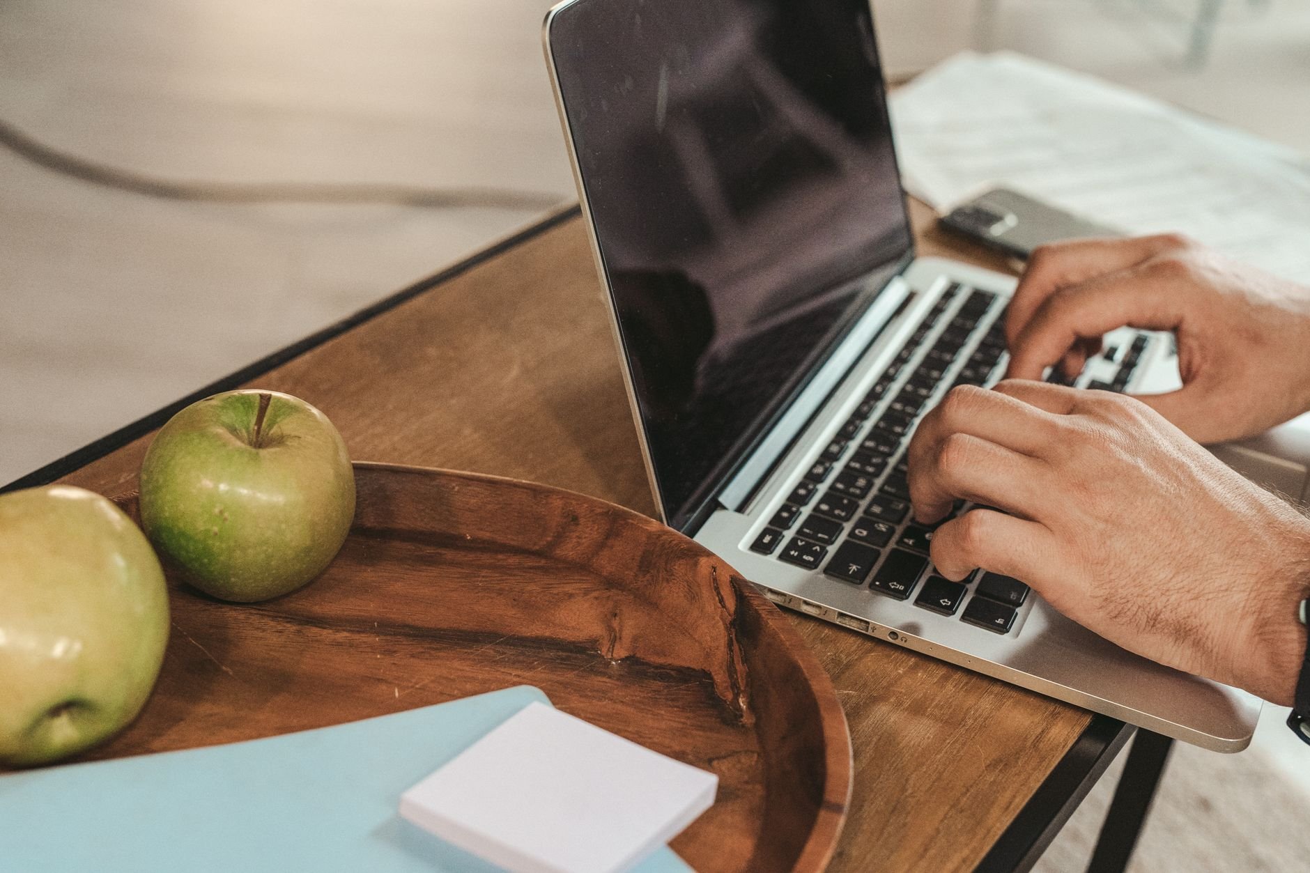 Hands typing on a laptop with green apples and sticky notes on a wooden desk, creating a productive work envir
